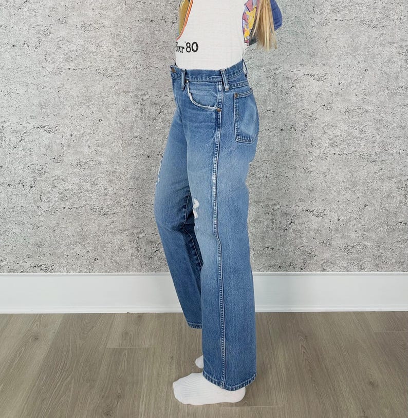 K&ouml;nnte beinhalten: Hellblaue Jeans mit geradem Bein. Die Jeans haben ein klassisches F&uuml;nf-Taschen-Design, einen Knopf- und Rei&szlig;verschluss sowie Used-Details. Die Jeans werden mit einem wei&szlig;en T-Shirt mit der Aufschrift "80" getragen.