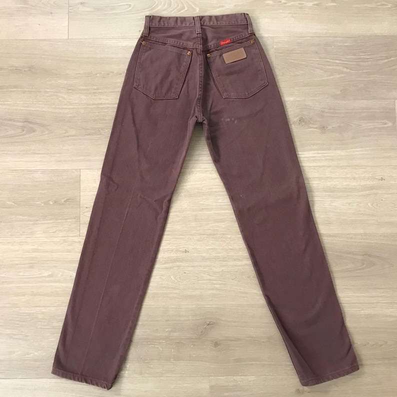 Op de afbeelding: Een paar mauve Wrangler-jeans, gezien vanaf de achterkant. De jeans hebben een klassiek ontwerp met vijf zakken, messing klinknagels en een leren patch. Het Wrangler-logo is zichtbaar op de achterzak.