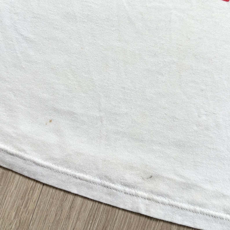 May include: A close-up of a white t-shirt with visible stains and a stitched hem. The fabric appears slightly wrinkled, and the shirt is lying on a wooden surface. The stains are brown and scattered across the shirt.