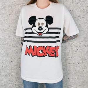 May include: White t-shirt featuring a Mickey Mouse graphic. The design includes Mickey's face above black and white horizontal stripes, with the name "MICKEY" in red block letters below. A classic and colorful design.