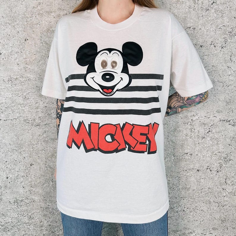 May include: White t-shirt featuring a Mickey Mouse graphic. The design includes Mickey's face above black and white horizontal stripes, with the name "MICKEY" in red block letters below. A classic and colorful design.
