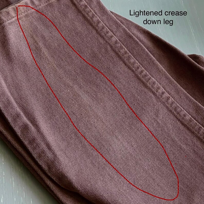 Op de afbeelding: Close-up van een deel van een bruine broek, met de textuur van de stof en de stikdetails. De tekst "Lightened crease down leg" is zichtbaar. De broek heeft een lichtere plek langs de pijp.
