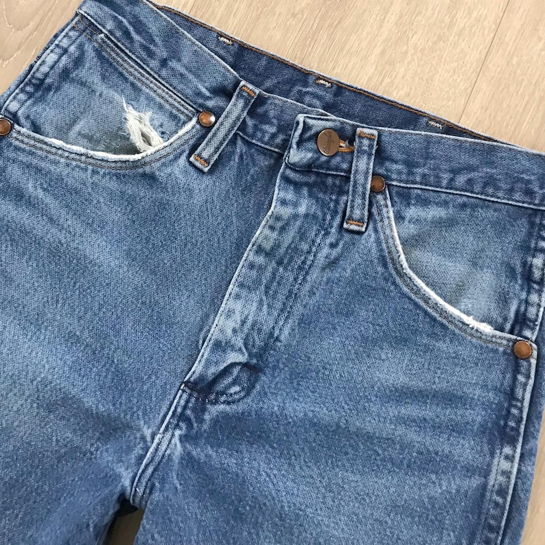 May include: A pair of blue denim jeans featuring a high-waisted design with a button fly closure, copper-toned metal rivets, and visible fraying along the edges of the front pockets. The fabric displays a classic medium-wash blue texture.