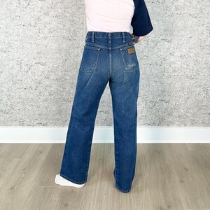 May include: Dark wash denim jeans with a high waist and a wide leg. The jeans have classic five-pocket styling and a brown leather patch on the back. The model is wearing a pink and navy blue baseball tee.