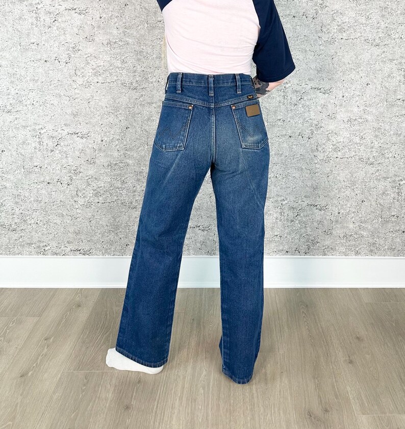 May include: Dark wash denim jeans with a high waist and a wide leg. The jeans have classic five-pocket styling and a brown leather patch on the back. The model is wearing a pink and navy blue baseball tee.
