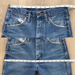 May include: Blue denim jeans featuring a button fly, copper-coloured rivets, and distressed pocket edges. A white measuring tape is positioned across the waist, showing a width of 35.5 cm, and across the hips, showing a width of 43 cm.