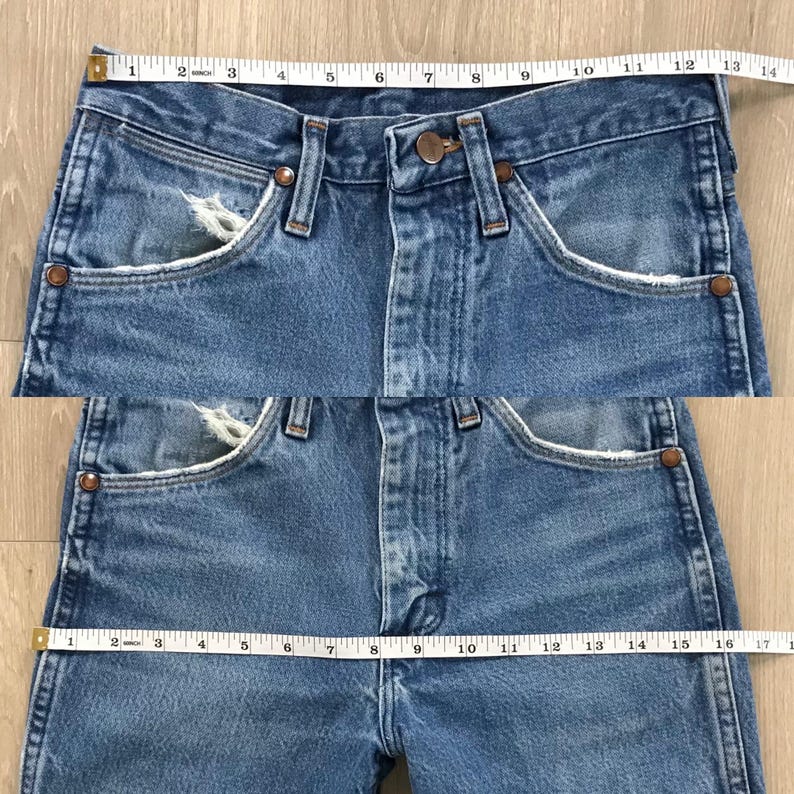 May include: Blue denim jeans featuring a button fly, copper-coloured rivets, and distressed pocket edges. A white measuring tape is positioned across the waist, showing a width of 35.5 cm, and across the hips, showing a width of 43 cm.