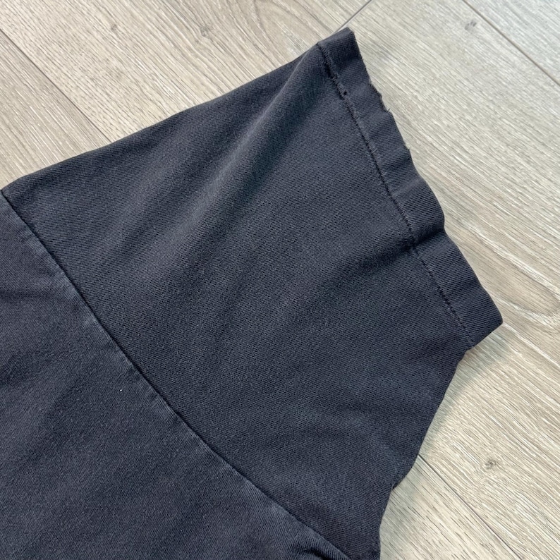 Peut inclure: Gros plan sur une manche de t-shirt gris fonc&eacute;. Le tissu semble l&eacute;g&egrave;rement us&eacute;, avec des coutures visibles et un bord pli&eacute;. La manche est inclin&eacute;e, montrant la texture du mat&eacute;riau.
