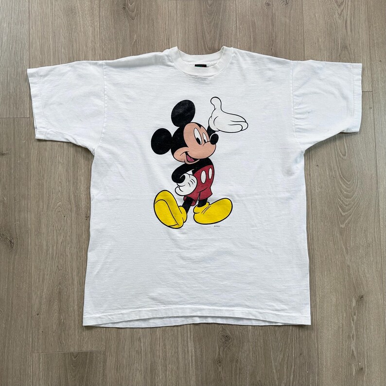 Op de afbeelding: Wit T-shirt met een Mickey Mouse-afbeelding. Het stripfiguur wordt afgebeeld met zwarte oren, een rode broek, gele schoenen en witte handschoenen. Het shirt is gemaakt van een zacht materiaal en heeft korte mouwen.