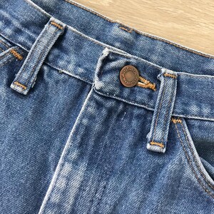 Op de afbeelding: Close-up van een blauwe spijkerbroek, met de tailleband, riemlussen en een koperkleurige knoop. De jeans heeft een klassiek ontwerp met zichtbare stiksels en een vervaagde uitstraling.