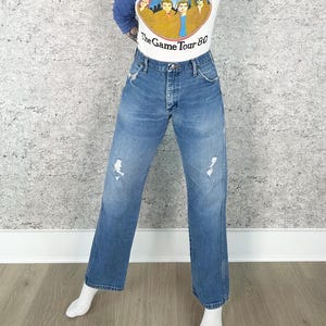 K&ouml;nnte beinhalten: Blaue Jeans aus Denim mit Used-Details. Die Jeans haben eine gerade Beinpassform und ein klassisches F&uuml;nf-Taschen-Design. Die Jeans werden mit einem wei&szlig;en Grafik-T-Shirt mit dem Text "The Game Tour 80" getragen.