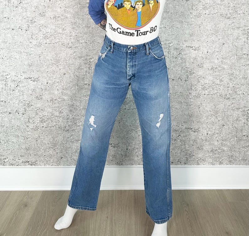 K&ouml;nnte beinhalten: Blaue Jeans aus Denim mit Used-Details. Die Jeans haben eine gerade Beinpassform und ein klassisches F&uuml;nf-Taschen-Design. Die Jeans werden mit einem wei&szlig;en Grafik-T-Shirt mit dem Text "The Game Tour 80" getragen.