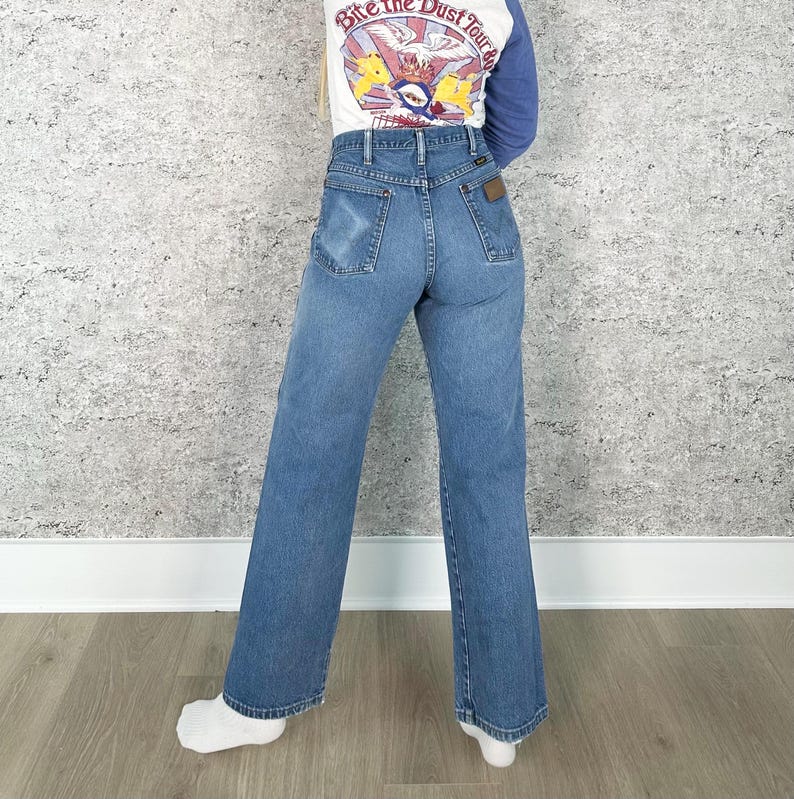 K&ouml;nnte beinhalten: Hellblaue Jeans mit hoher Taille und weitem Bein. Die Jeans haben ein klassisches F&uuml;nf-Taschen-Design und einen braunen Lederaufn&auml;her auf der R&uuml;ckseite. Die Person tr&auml;gt ein wei&szlig;es und blaues Langarmshirt mit dem Text "Bite the Dust Tour".