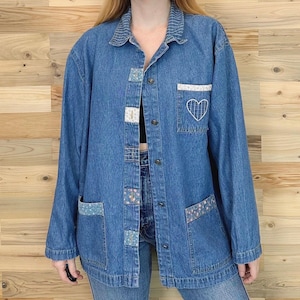 May include: A medium-wash denim jacket with long sleeves, a collared neckline, and button closures. The jacket features decorative patches and a white embroidered heart on the pocket. The jacket has two front pockets and a relaxed fit.