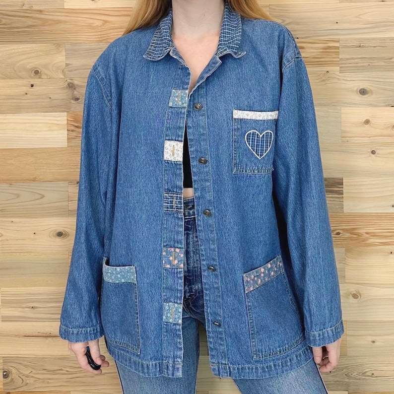 May include: A medium-wash denim jacket with long sleeves, a collared neckline, and button closures. The jacket features decorative patches and a white embroidered heart on the pocket. The jacket has two front pockets and a relaxed fit.