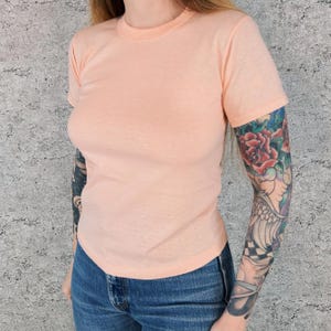Puede incluir: Una camiseta melocotón de manga corta con cuello redondo. La camiseta está hecha de un material suave y ligero. La persona lleva vaqueros azules. El fondo es una pared gris texturizada.