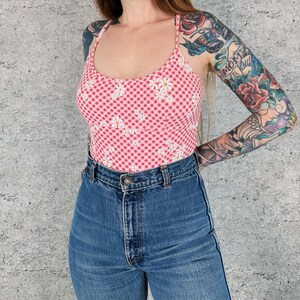 May include: A pink and white gingham print tank top with a daisy floral pattern. The top is paired with high-waisted blue denim jeans. The tank top has a scoop neck and spaghetti straps.