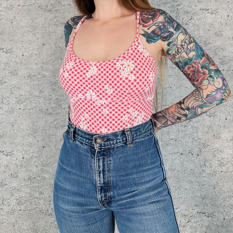 May include: A pink and white gingham print tank top with a daisy floral pattern. The top is paired with high-waisted blue denim jeans. The tank top has a scoop neck and spaghetti straps.