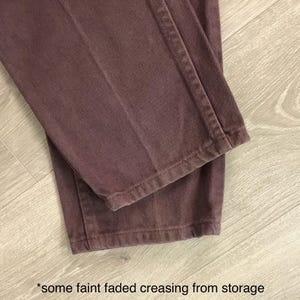 Op de afbeelding: Close-up van een mauvekleurige broek. De stof vertoont vage, vervaagde kreukels van opslag. De broek is opgevouwen, waardoor de zoom en naden zichtbaar zijn. De achtergrond is een lichtgekleurd houten oppervlak.