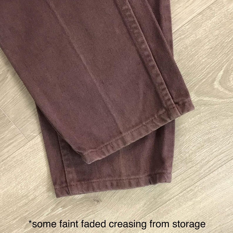 Op de afbeelding: Close-up van een mauvekleurige broek. De stof vertoont vage, vervaagde kreukels van opslag. De broek is opgevouwen, waardoor de zoom en naden zichtbaar zijn. De achtergrond is een lichtgekleurd houten oppervlak.