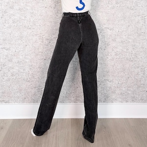 Pu&ograve; includere: Jeans neri in denim a vita alta e gamba dritta. I jeans hanno un classico design a cinque tasche e una chiusura con bottone e cerniera. La modella indossa una maglietta bianca con una grafica blu.