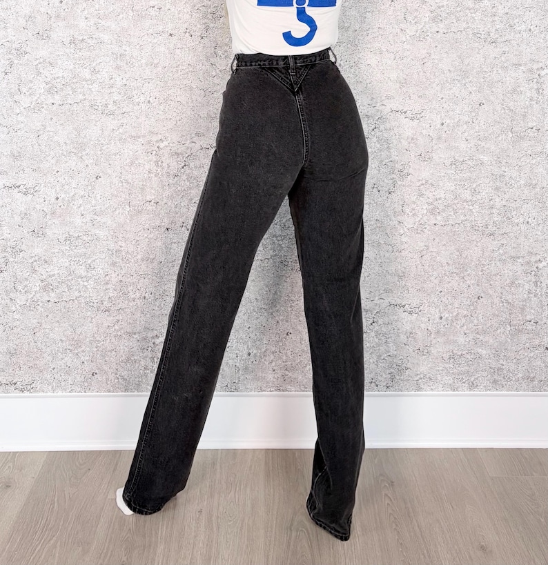 Pu&ograve; includere: Jeans neri in denim a vita alta e gamba dritta. I jeans hanno un classico design a cinque tasche e una chiusura con bottone e cerniera. La modella indossa una maglietta bianca con una grafica blu.
