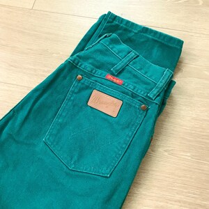 May include: Teal-colored Wrangler jeans with a classic five-pocket design. The jeans feature a brown leather patch with the Wrangler logo on the back pocket. The back pocket has a stitched design. The jeans are folded on a light wood surface.