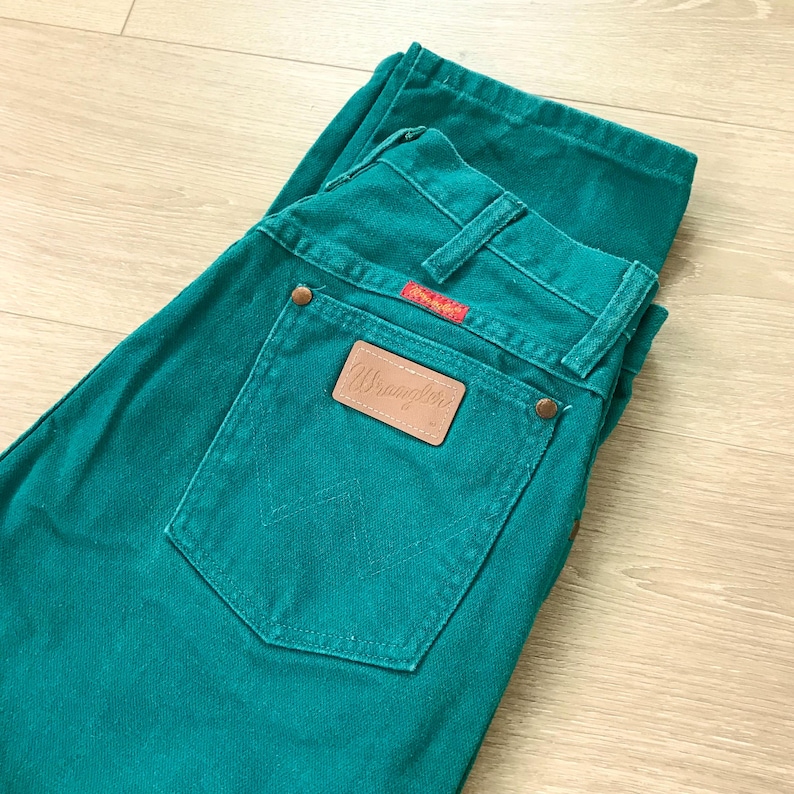 May include: Teal-colored Wrangler jeans with a classic five-pocket design. The jeans feature a brown leather patch with the Wrangler logo on the back pocket. The back pocket has a stitched design. The jeans are folded on a light wood surface.