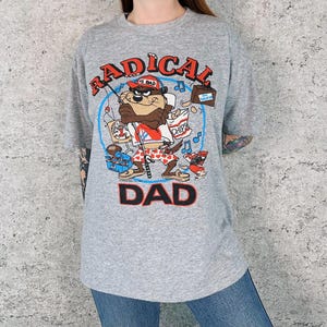 Pode incluir: Uma camiseta cinza com o personagem de desenho animado Taz e as palavras "RADICAL DAD" em vermelho e preto. O gráfico inclui uma vara de pescar, batatas fritas e um boombox, criando um visual divertido e vintage.