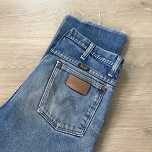 May include: Pale blue denim jeans with a classic fit. The jeans feature a brown leather Wrangler patch on the back pocket and the Wrangler logo on the back pocket and waistband. The jeans have a faded wash and a raw hem.