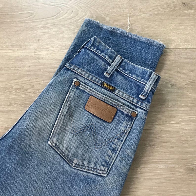May include: Pale blue denim jeans with a classic fit. The jeans feature a brown leather Wrangler patch on the back pocket and the Wrangler logo on the back pocket and waistband. The jeans have a faded wash and a raw hem.