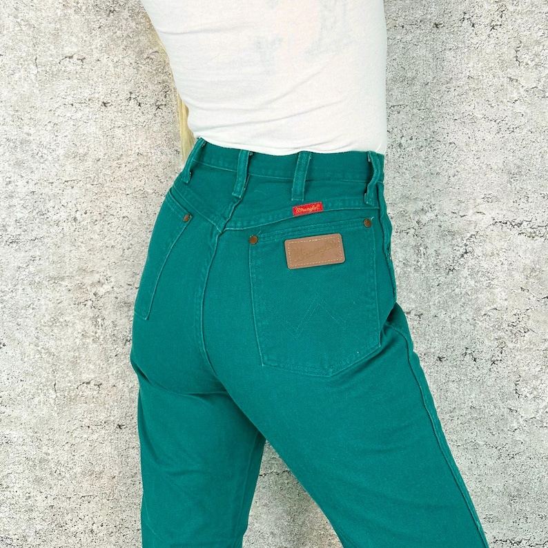 May include: Teal-colored denim jeans with a high waist and a brown leather patch on the back pocket. The jeans have a classic five-pocket design and a red tag. The person is wearing a white top.