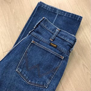 May include: Dark blue denim jeans with a classic five-pocket design. The jeans feature a straight leg cut and a Wrangler brand label on the back pocket. The stitching is a contrasting orange-brown color.