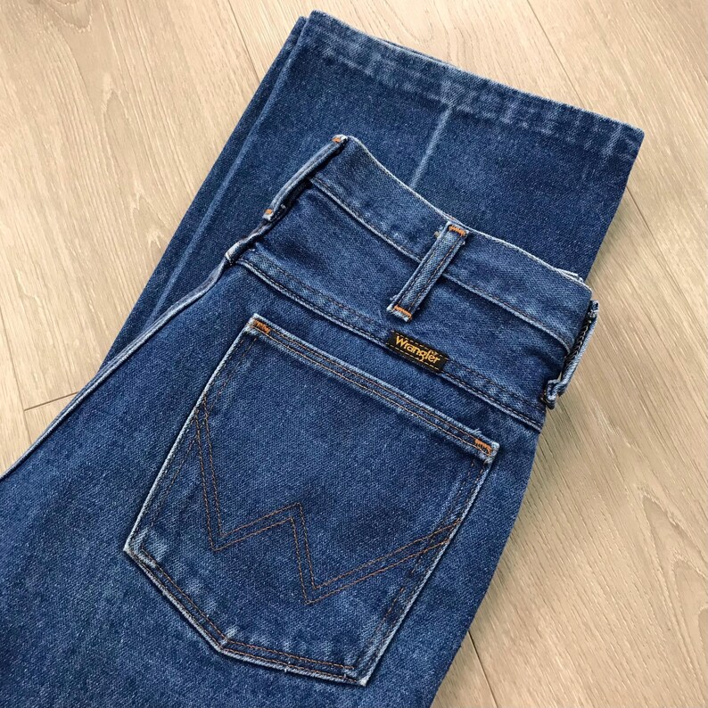 May include: Dark blue denim jeans with a classic five-pocket design. The jeans feature a straight leg cut and a Wrangler brand label on the back pocket. The stitching is a contrasting orange-brown color.