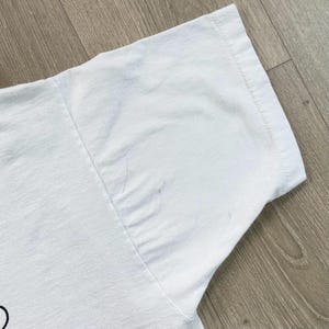 Op de afbeelding: Close-up van een witte t-shirtmouw. De mouw is gemaakt van een zachte, witte stof en heeft een gestikte zoom. Er zitten een paar kleine vlekken op de stof. Het shirt ligt op een houten ondergrond.