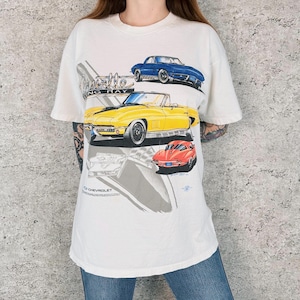 May include: White t-shirt with illustrations of classic Chevrolet Corvette Sting Ray cars in blue, yellow, and red. The shirt includes the Chevrolet logo and the words "Corvette Sting Ray".