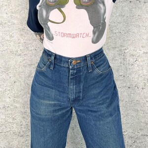 May include: A person wearing a white t-shirt with a graphic and the text "..STORMWATCH.." and blue denim jeans. The jeans have a button closure and classic five-pocket design. The t-shirt has navy blue sleeves.