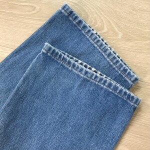 Op de afbeelding: Close-up van twee lichtblauwe denim jeanszomen. De zomen zijn omgeslagen, waardoor de stiksels en de textuur van de denimstof zichtbaar worden. De jeans liggen op een lichtbruin houten oppervlak.