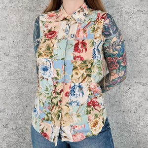 Può includere: Una camicia senza maniche con bottoni caratterizzata da un design patchwork con vari motivi floreali nei toni del rosso, blu, rosa e verde su sfondo crema. La camicia presenta un colletto classico e una chiusura frontale con bottoni.