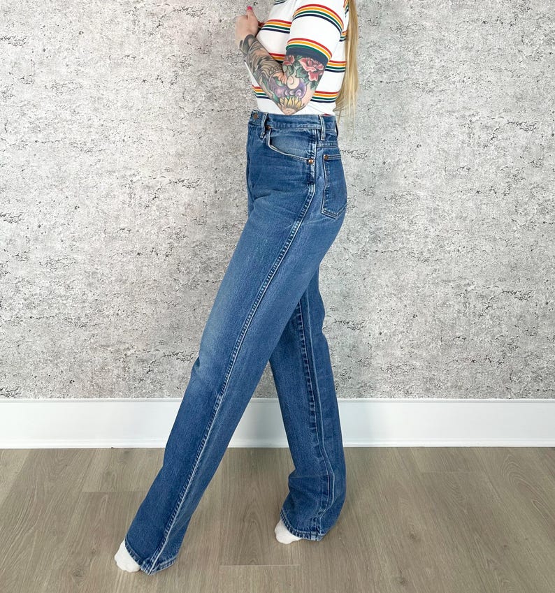 Puede incluir: Vaqueros de mezclilla azul de lavado medio, de cintura alta. Los vaqueros tienen un dise&ntilde;o cl&aacute;sico de cinco bolsillos y un corte de pierna recta. La modelo lleva una camiseta de manga corta a rayas con rayas arco&iacute;ris.
