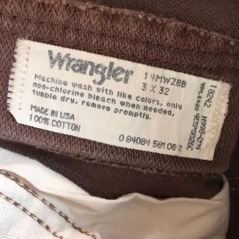 Op de afbeelding: Close-up van een bruine Wrangler-jeans. Het label toont de merknaam en wasvoorschriften. Maat 3 x 32. De jeans is gemaakt van 100% katoen en gemaakt in de VS.