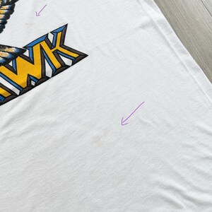 May include: White t-shirt with a large graphic design on the upper left. The graphic features the word "HAWK" in yellow and blue block letters, with a stylized eagle wing design. The shirt is slightly wrinkled.