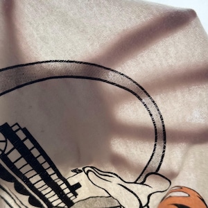 May include: Close-up of a light pink fabric with a black and white cartoon graphic. The graphic includes a large black circle, a building, and a cartoon hand. The fabric is slightly wrinkled.