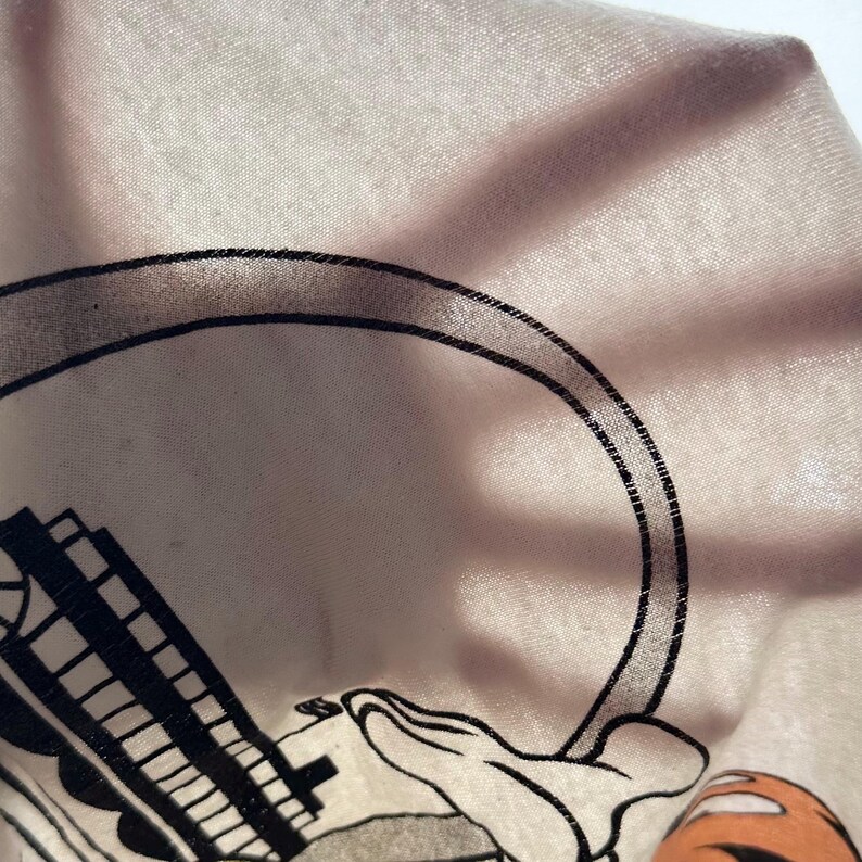 May include: Close-up of a light pink fabric with a black and white cartoon graphic. The graphic includes a large black circle, a building, and a cartoon hand. The fabric is slightly wrinkled.