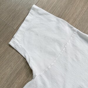Op de afbeelding: Close-up van een wit T-shirt, met de schouder en het bovenste borstgedeelte. De stof lijkt een zacht katoen te zijn, met zichtbare naden en een licht getextureerd oppervlak. Het shirt is schoon en lijkt ongedragen.