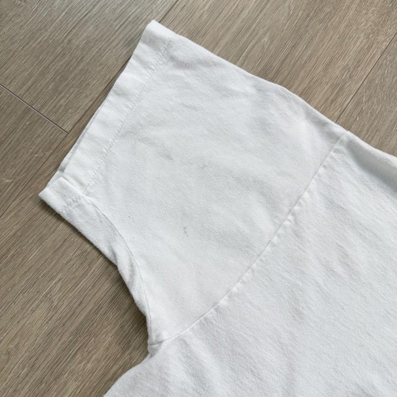 Op de afbeelding: Close-up van een wit T-shirt, met de schouder en het bovenste borstgedeelte. De stof lijkt een zacht katoen te zijn, met zichtbare naden en een licht getextureerd oppervlak. Het shirt is schoon en lijkt ongedragen.