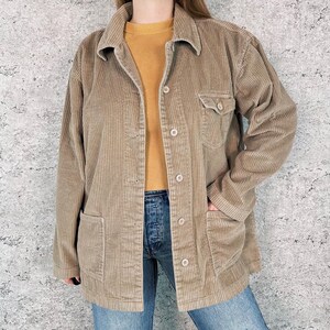 May include: A tan corduroy jacket with a collared neckline, button-down front, and two front pockets. The jacket is worn over a mustard-colored sweater and blue jeans. The jacket has long sleeves and a relaxed fit.
