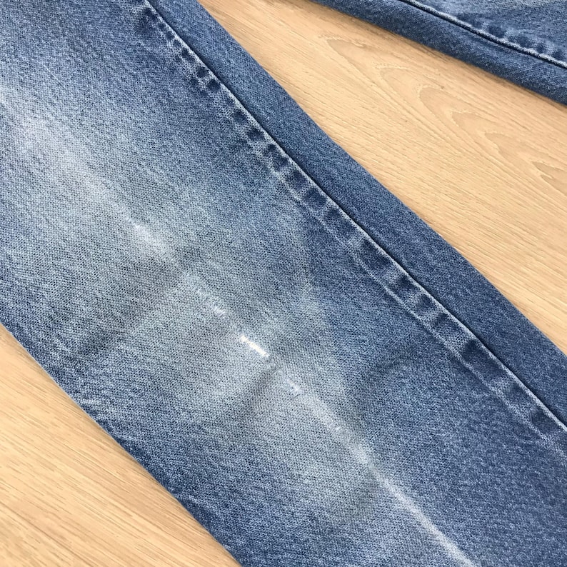 May include: A pair of blue denim jeans featuring a classic five-pocket construction with visible stitching along the seams. The fabric displays a faded wash effect on the thigh area, highlighting the texture of the cotton denim material.