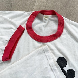 May include: White t-shirt with a red collar and sleeve trim. The shirt features a black graphic on the lower right side. The fabric appears soft and well-worn, suggesting a vintage style.