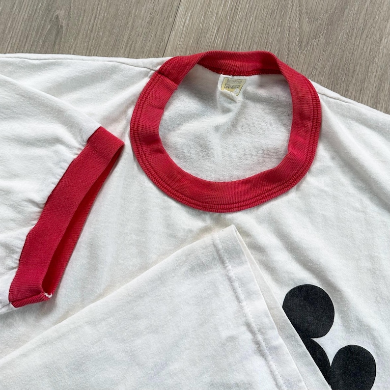 May include: White t-shirt with a red collar and sleeve trim. The shirt features a black graphic on the lower right side. The fabric appears soft and well-worn, suggesting a vintage style.